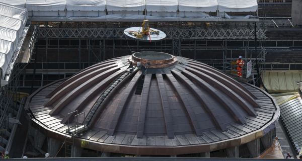 Workers in safety gear are working on the copper roof of a large dome structure surrounded by scaffolding, with a crane lifting a section of the roof.