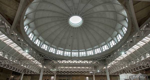 A large, empty industrial building under renovation with a circular dome ceiling, metal supports, and construction materials scattered around. Orange barriers cordon off parts of the site.